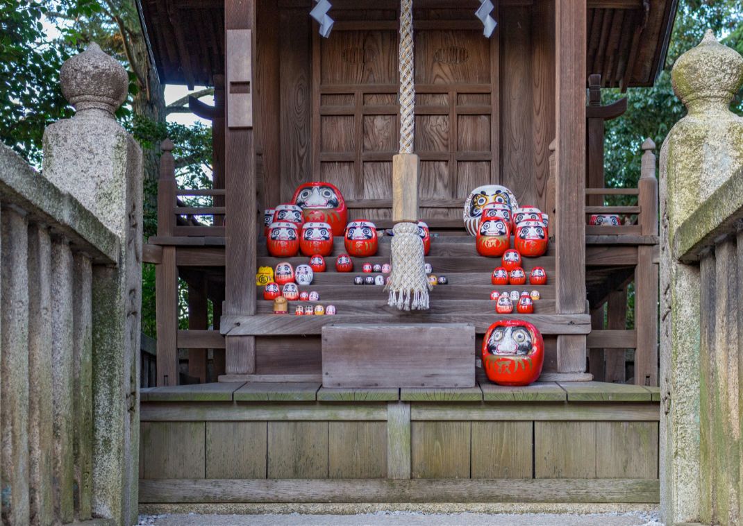 Vista panorámica de la entrada de un santuario con numerosos muñecos Daruma de diferentes tamaños colocados en los escalones de madera.