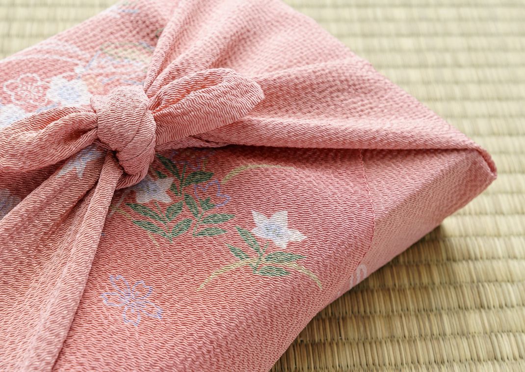 Un pañuelo japonés tradicional furoshiki, de color rosa con diseños florales, cuidadosamente atado para envolver una caja rectangular, descansando sobre un tatami.