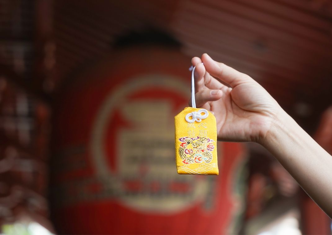 Una mano sostiene un omamori amarillo bordado, un amuleto japonés de buena suerte, con una gran linterna roja desenfocada al fondo.