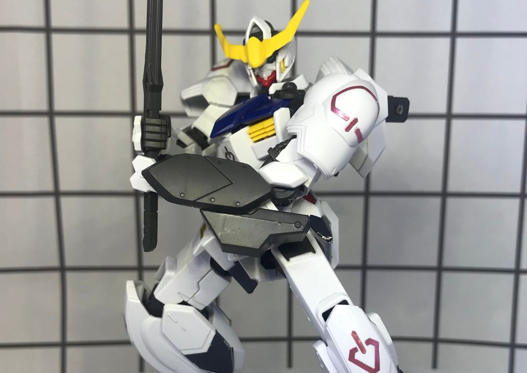 Primer plano de una figura de maqueta Gundam en blanco, azul y amarillo, posada frente a azulejos cuadrados.