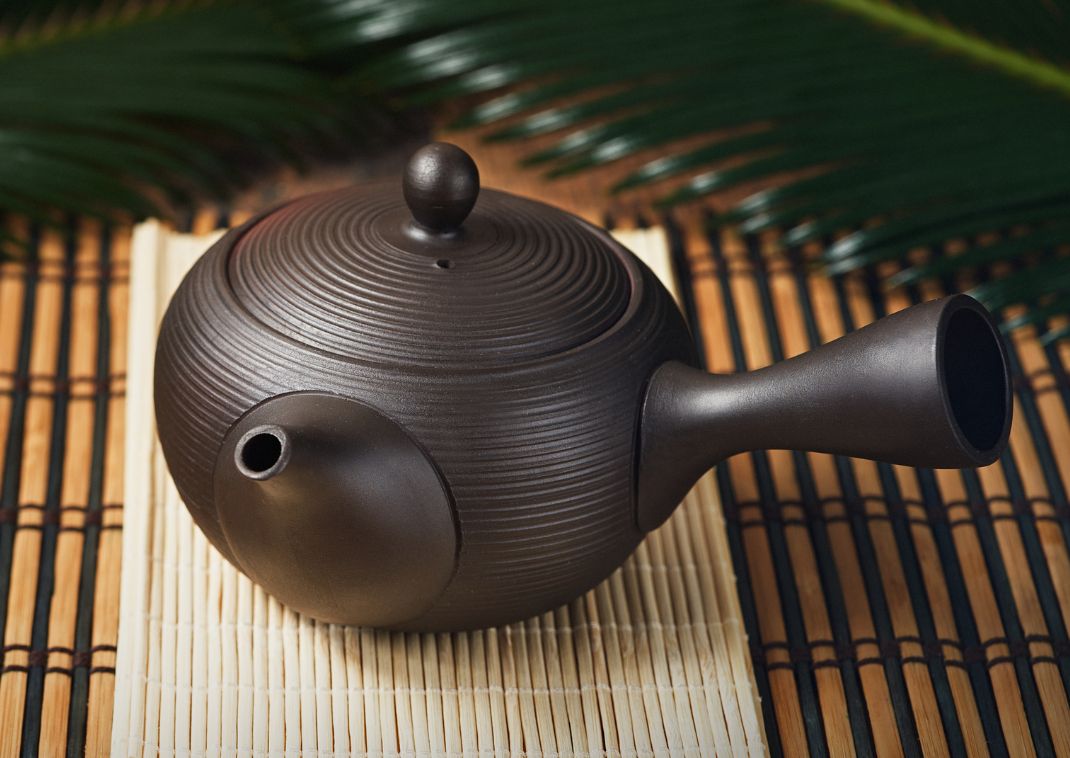 Una tetera japonesa tradicional de cerámica negra con mango lateral, colocada sobre un tapete de bambú.