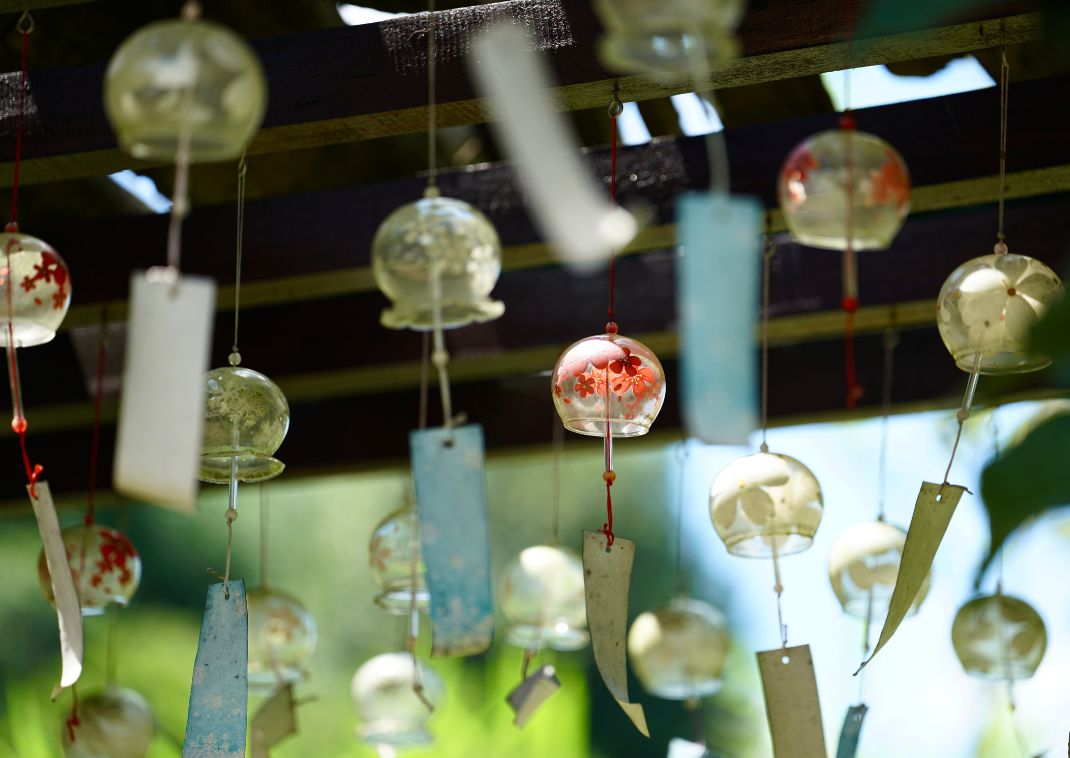 Varios carillones de viento de vidrio (furin) con diseños pintados y tiras de papel colgantes, suspendidos de vigas de madera al aire libre bajo la luz del sol.