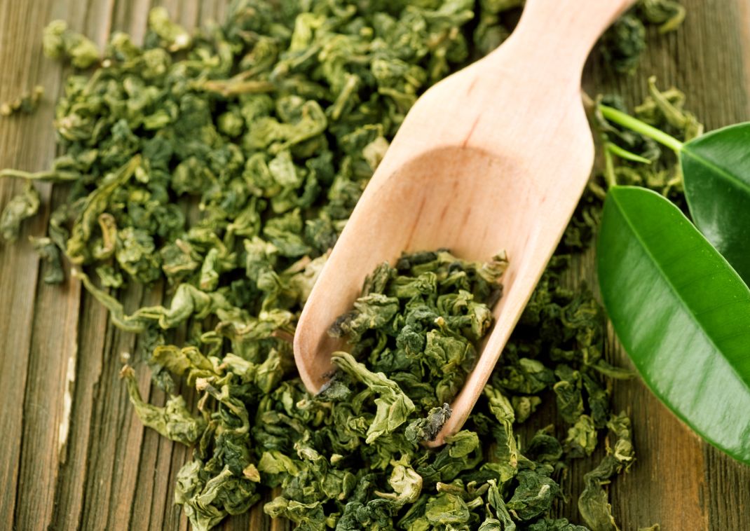 Hojas de té verde secas esparcidas sobre una superficie de madera, con una cuchara de madera y algunas hojas verdes.