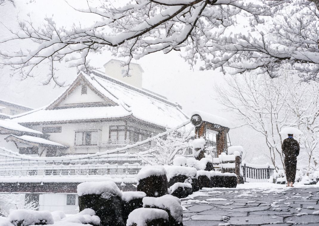 Persona con ropa tradicional caminando bajo intensa nevada cerca de edificios de estilo japonés y árboles cubiertos de nieve
