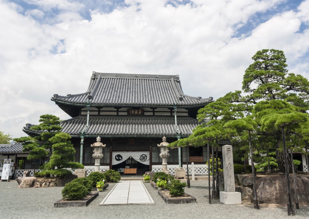 Edificio tradicional de templo japonés con techo de tejas, faroles de piedra y árboles cuidados en primer plano.