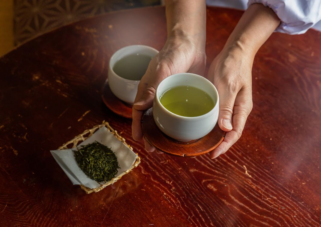 Persona sirviendo dos tazas de té verde sobre una mesa de madera, con té de hojas sueltas en una pequeña bandeja cercana.
