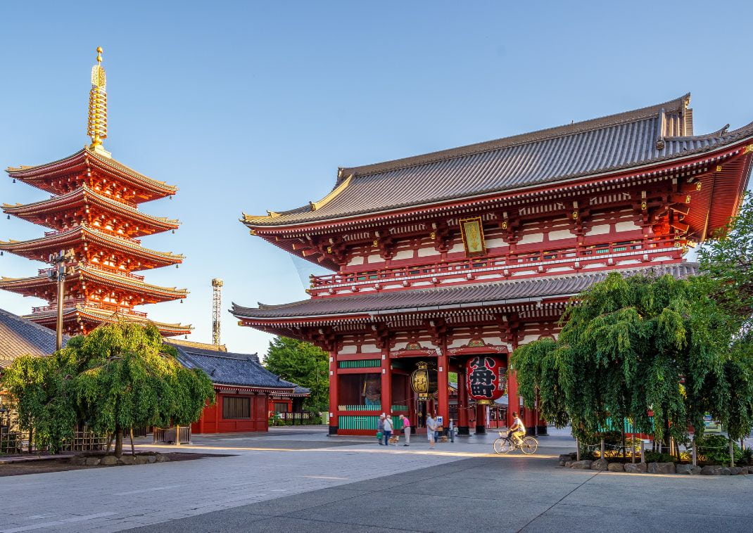 Gran puerta de templo budista roja y blanca y pagoda de cinco pisos, con personas caminando cerca y árboles en primer plano.