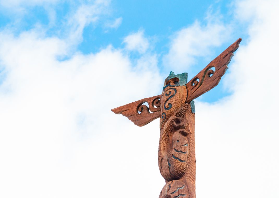 Primer plano de un tótem tradicional de madera con tallados intrincados, sobre un cielo azul con nubes