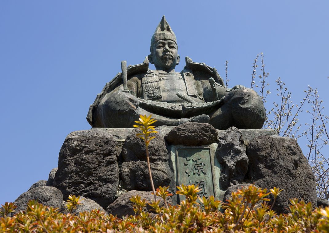 Estatua de bronce de un samurái sentado con armadura sobre un pedestal de piedra, con plantas en primer plano y cielo azul despejado