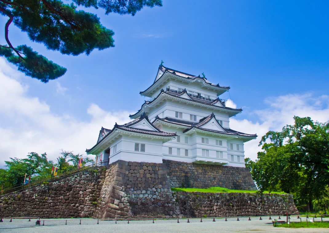 Castillo japonés tradicional con paredes blancas y techos de tejas, rodeado de muros de piedra y árboles bajo un cielo azul.
