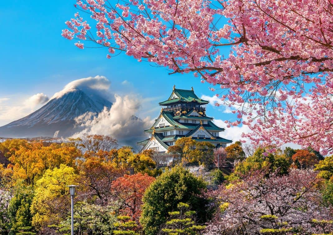 Castillo japonés tradicional rodeado de cerezos en flor y árboles coloridos, con el monte Fuji al fondo bajo un cielo azul.