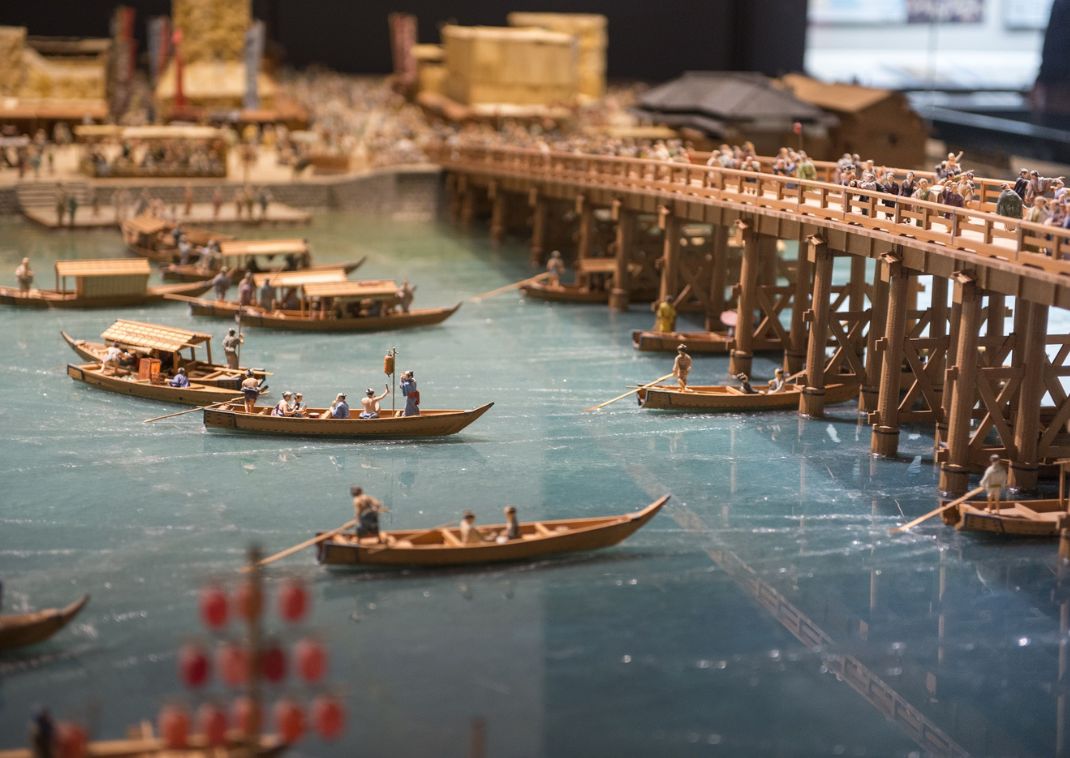 Modelo en miniatura detallado de barcos de madera y un puente sobre un río, con pequeñas figuras humanas que representan la vida histórica japonesa.