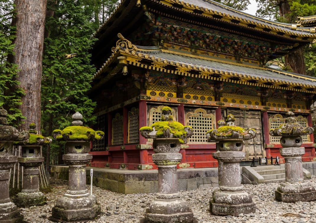 Edificio de templo japonés ornamentado con faroles de piedra cubiertos de musgo en un entorno boscoso.