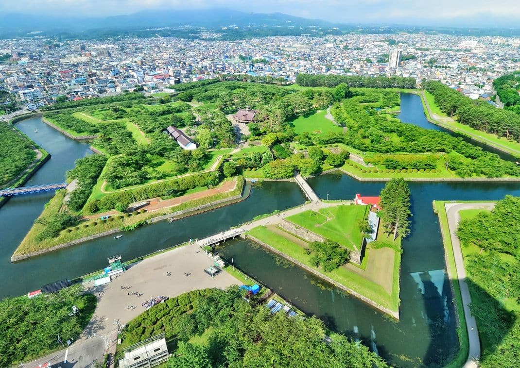 Vista aérea de una gran fortaleza en forma de estrella rodeada de árboles verdes y agua, con una ciudad y montañas al fondo.