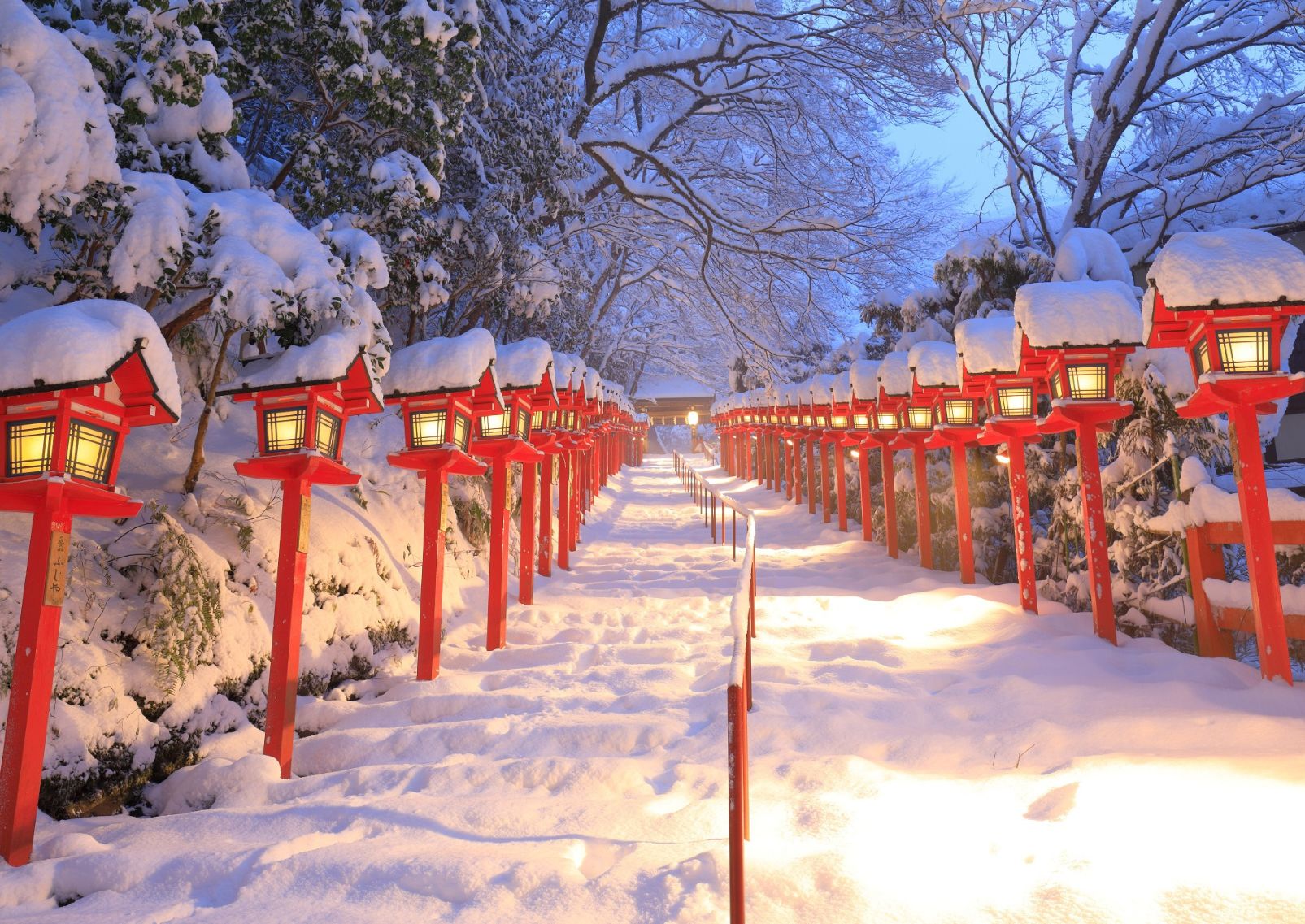 Escalera de piedra cubierta de nieve, flanqueada por faroles rojos tradicionales japoneses iluminados, rodeada de árboles nevados.