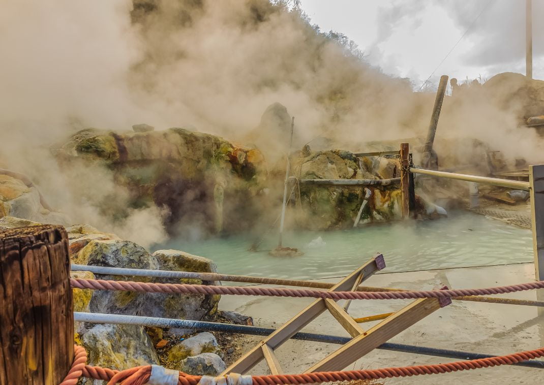 Piscina termal al aire libre con agua azul lechosa y vapor, rodeada de rocas y barandillas de madera en Hakone, Japón.