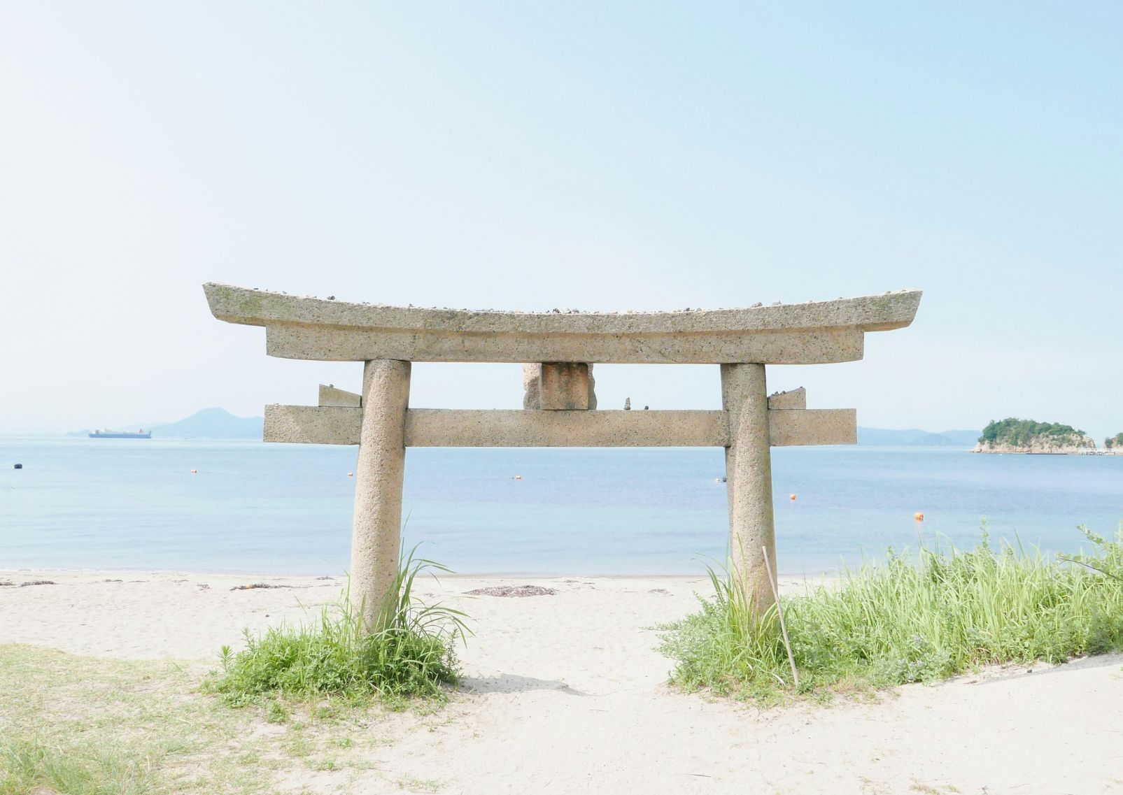 Una puerta torii de piedra medio sumergida en la arena con el mar al fondo