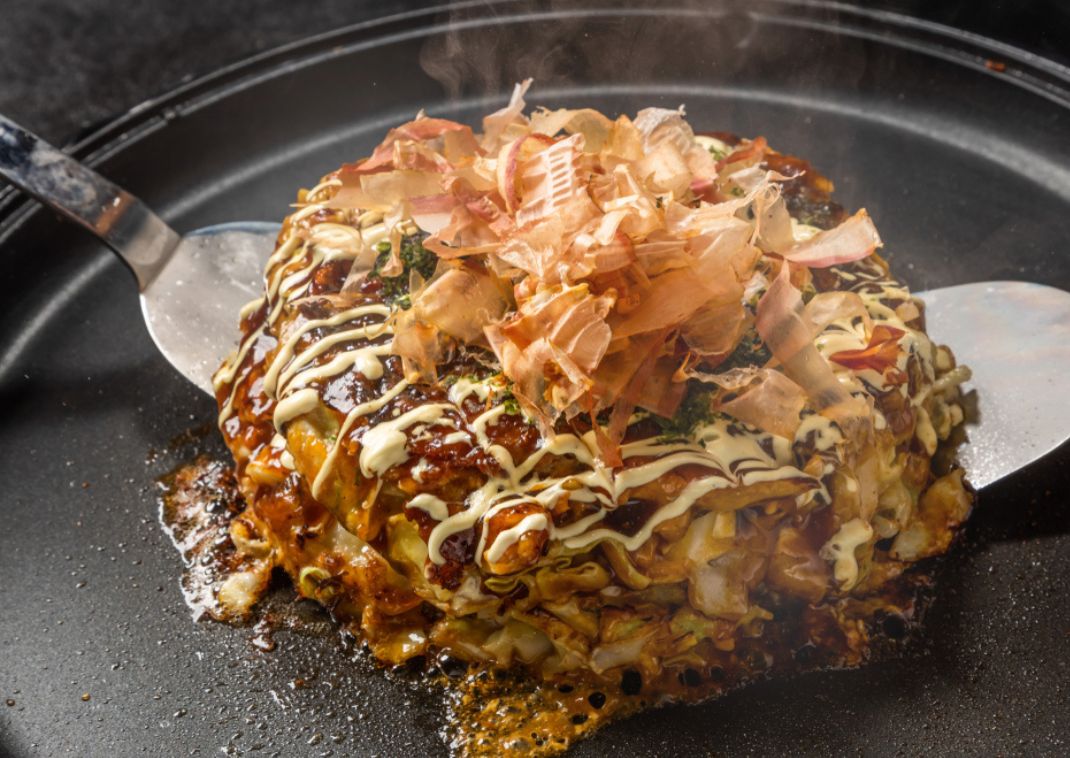 Okonomiyaki caliente, una tortilla japonesa salada cubierta con mayonesa, salsa okonomiyaki y copos de bonito, servida en una plancha caliente con espátulas.