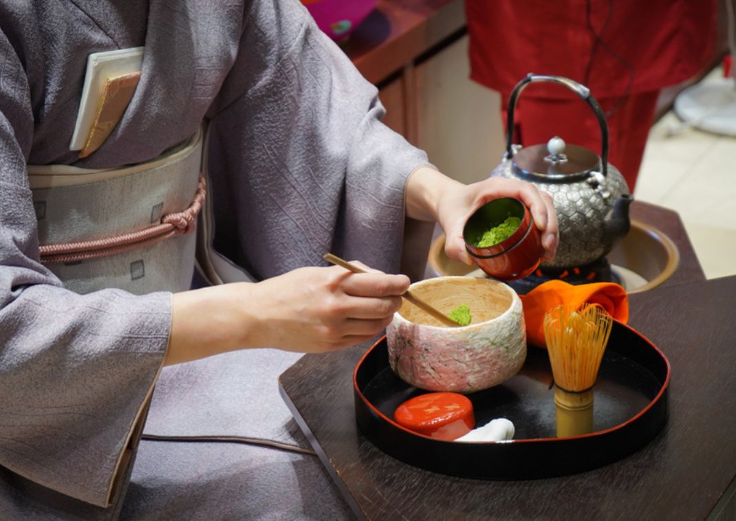 Persona con kimono lavanda agregando polvo de matcha a un cuenco de té con una cucharilla de bambú, con utensilios de té dispuestos en una bandeja.
