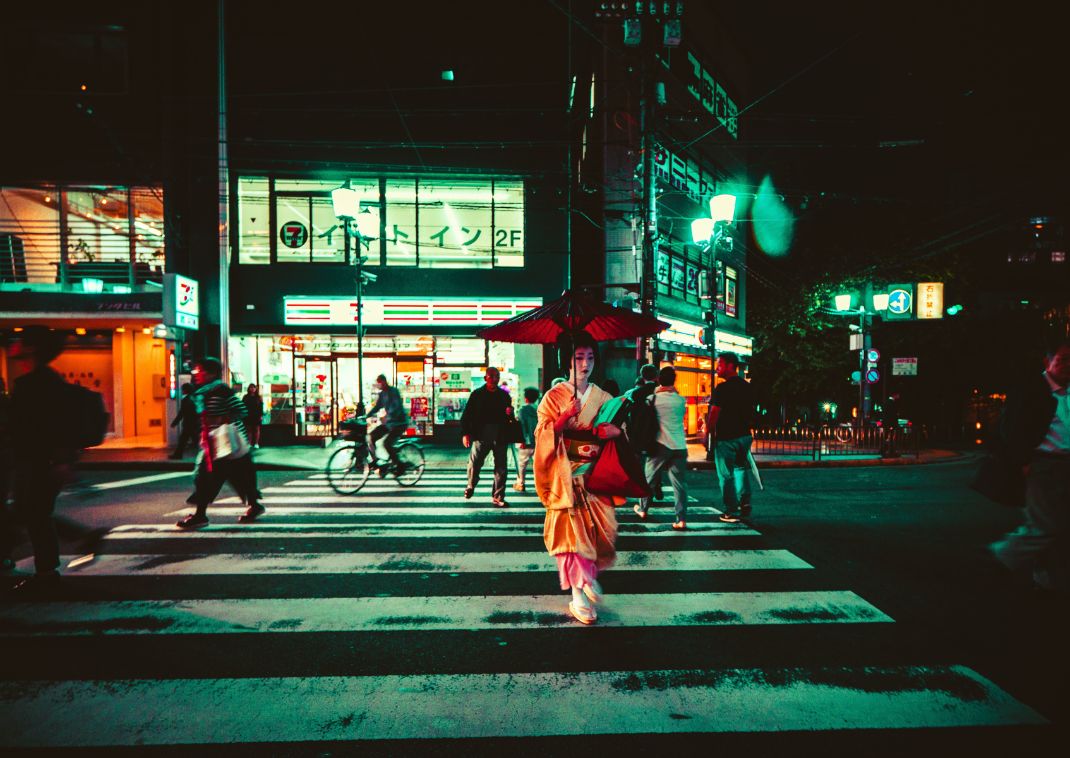 Persona con kimono y paraguas rojo cruzando una calle concurrida de la ciudad por la noche, con luces de neón y una tienda de conveniencia al fondo.