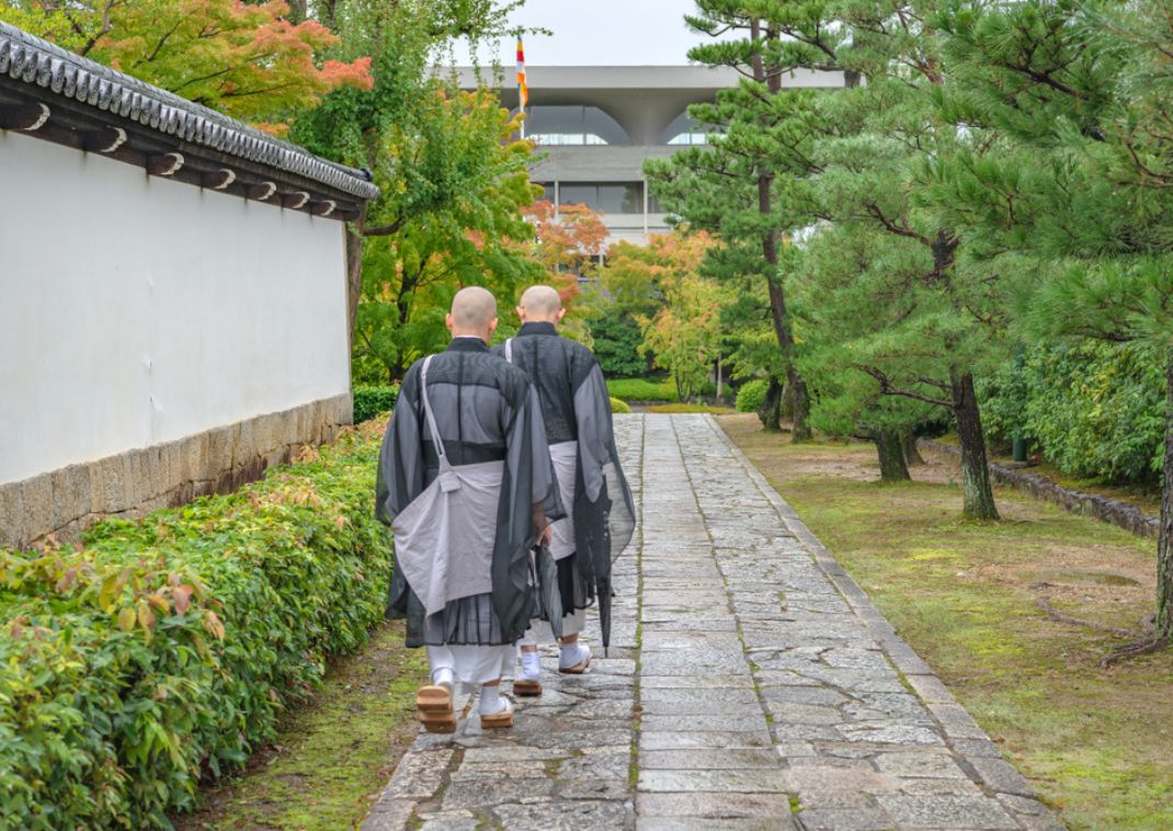  Dos monjes budistas con túnicas tradicionales caminando por un sendero de piedra en un jardín de templo japonés con pinos y follaje otoñal.