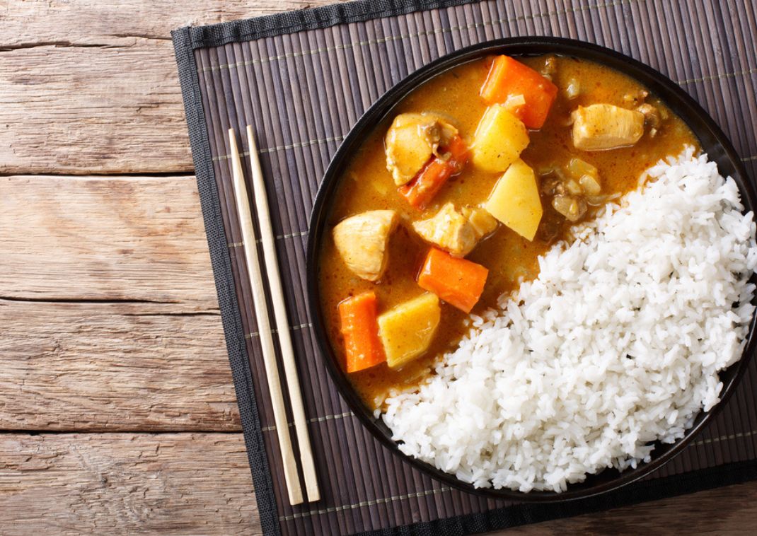 Curry japonés con arroz, trozos de pollo, patatas y zanahorias servido en un plato negro con arroz blanco y palillos sobre un mantel de bambú.