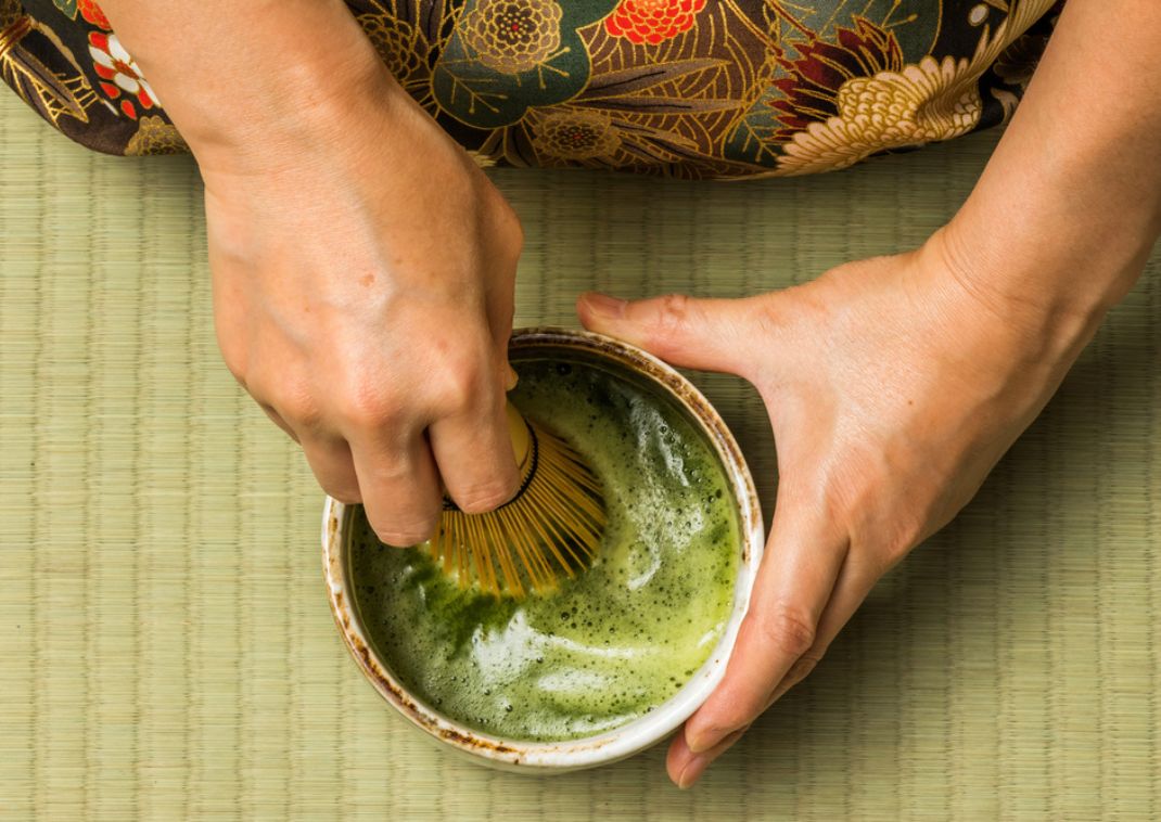 Primer plano de manos batiendo té verde matcha en un cuenco sobre tatami durante una ceremonia japonesa del té.