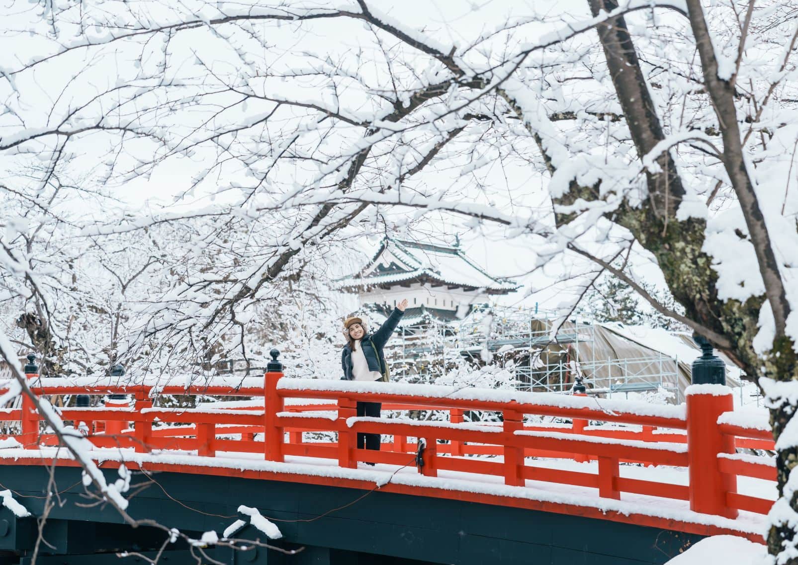 Puente rojo tradicional japonés cubierto de nieve, con árboles nevados y un castillo japonés al fondo.