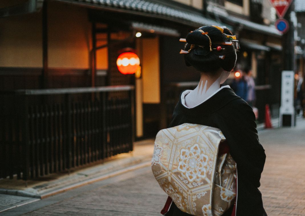 Geisha con kimono negro y obi ornamentado caminando por una calle tradicional japonesa al atardecer, vista desde atrás.