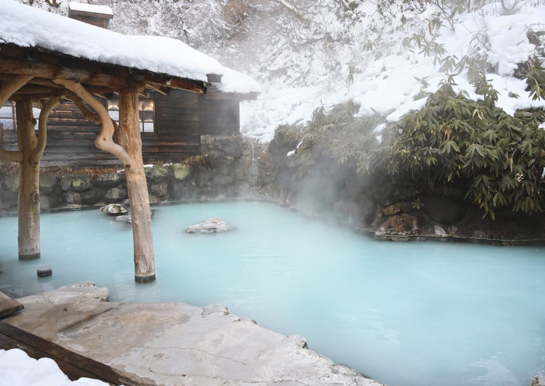 Onsen japonés al aire libre con agua azul lechosa, rodeado de rocas y estructuras de madera cubiertas de nieve en un paisaje invernal.