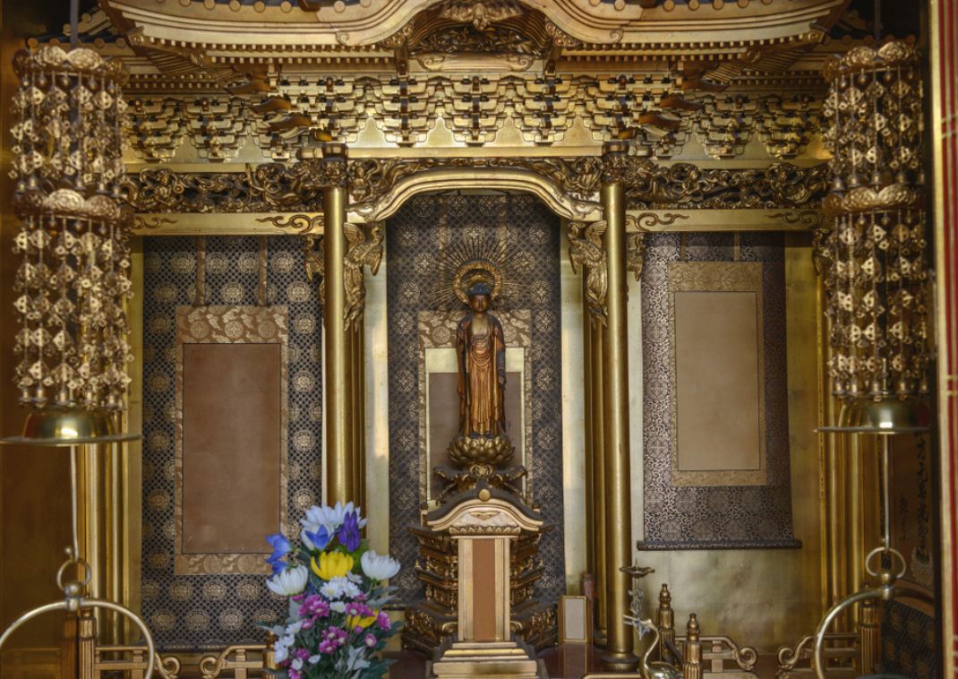Altar budista dorado ornamentado con una estatua de pie, decoraciones intrincadas y ofrendas de flores en el interior de un templo japonés.