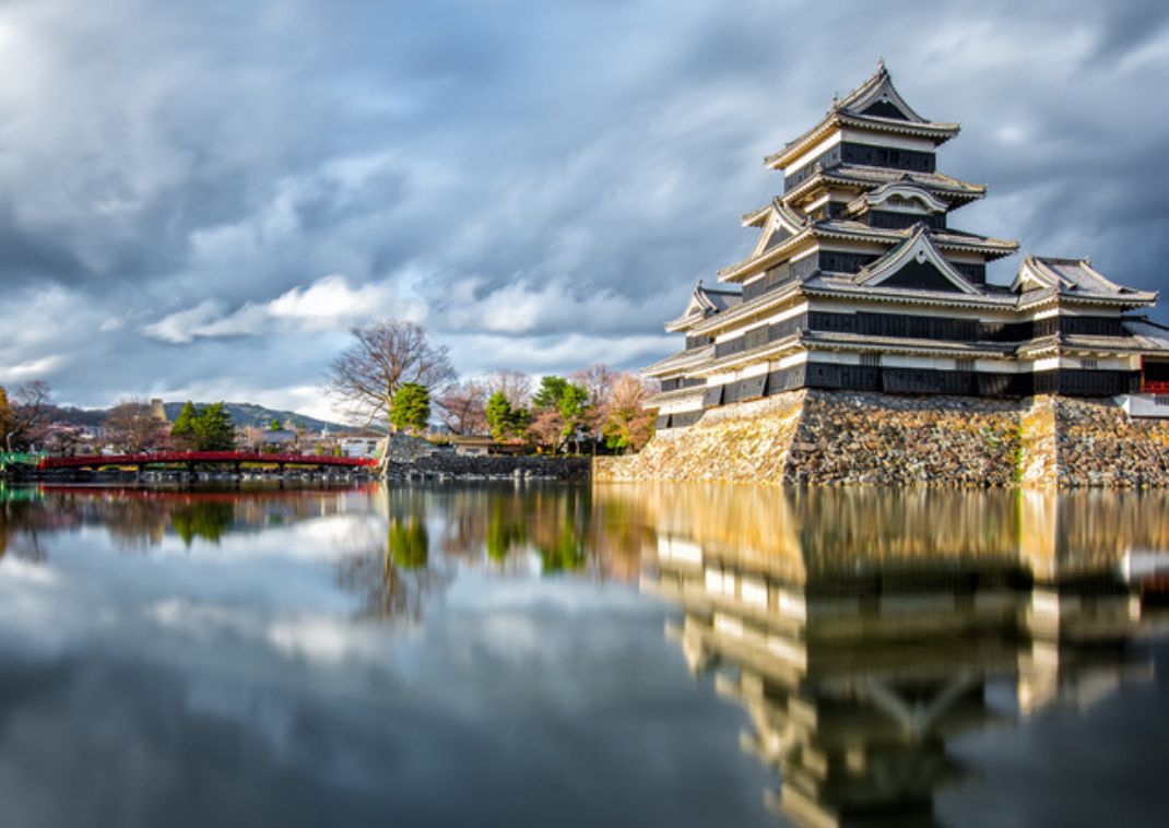 Castillo japonés histórico de paredes de madera negra reflejado en un foso tranquilo, con un puente rojo y árboles bajo un cielo nublado dramático.