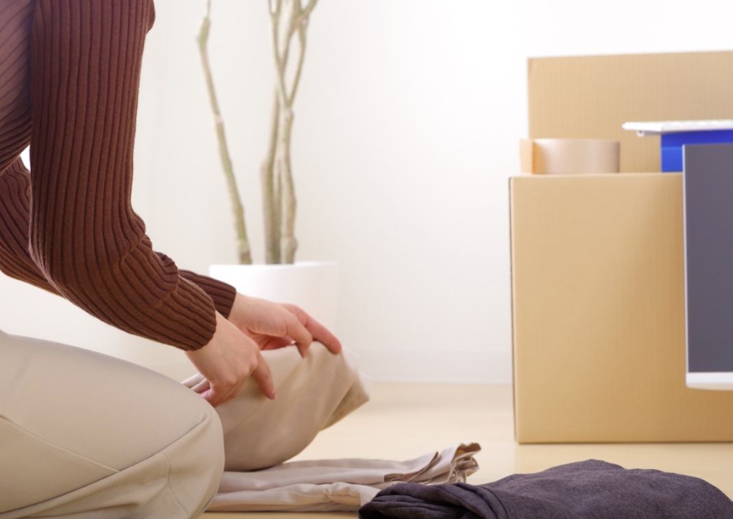 Persona arrodillada en el suelo doblando pantalones beige junto a una pila de ropa y cajas de mudanza de cartón.