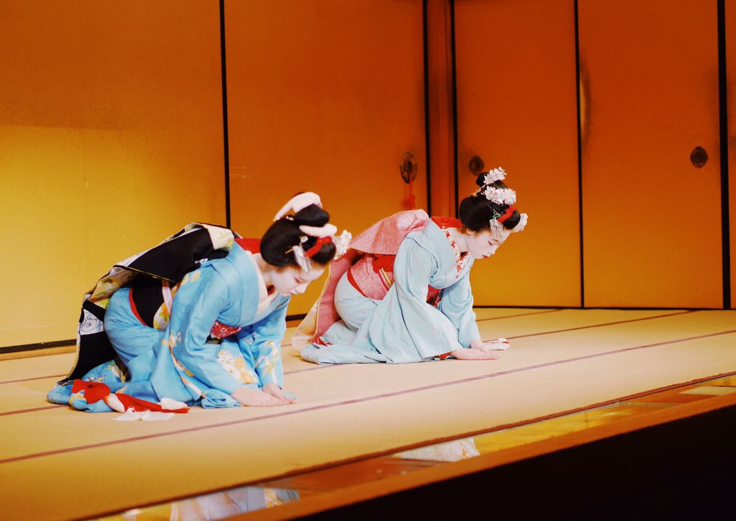 Dos geishas con kimonos coloridos realizando una reverencia tradicional sobre un escenario de tatami con un biombo dorado de fondo.