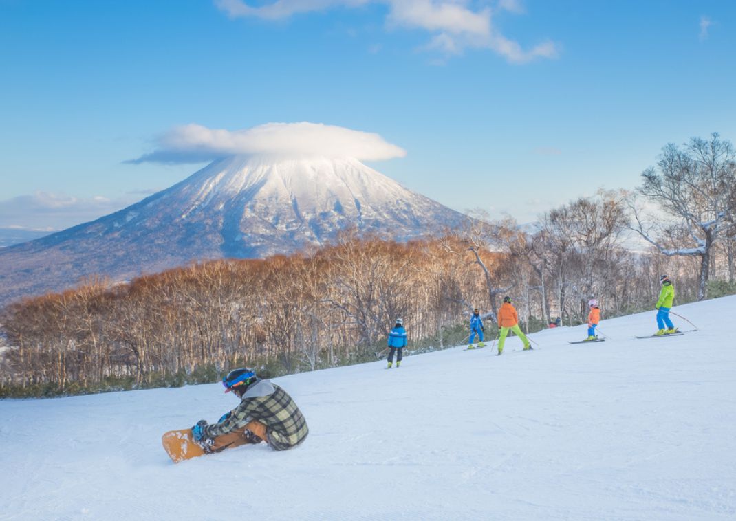 Esquiadores y snowboarder en una pista con árboles y un volcán nevado al fondo