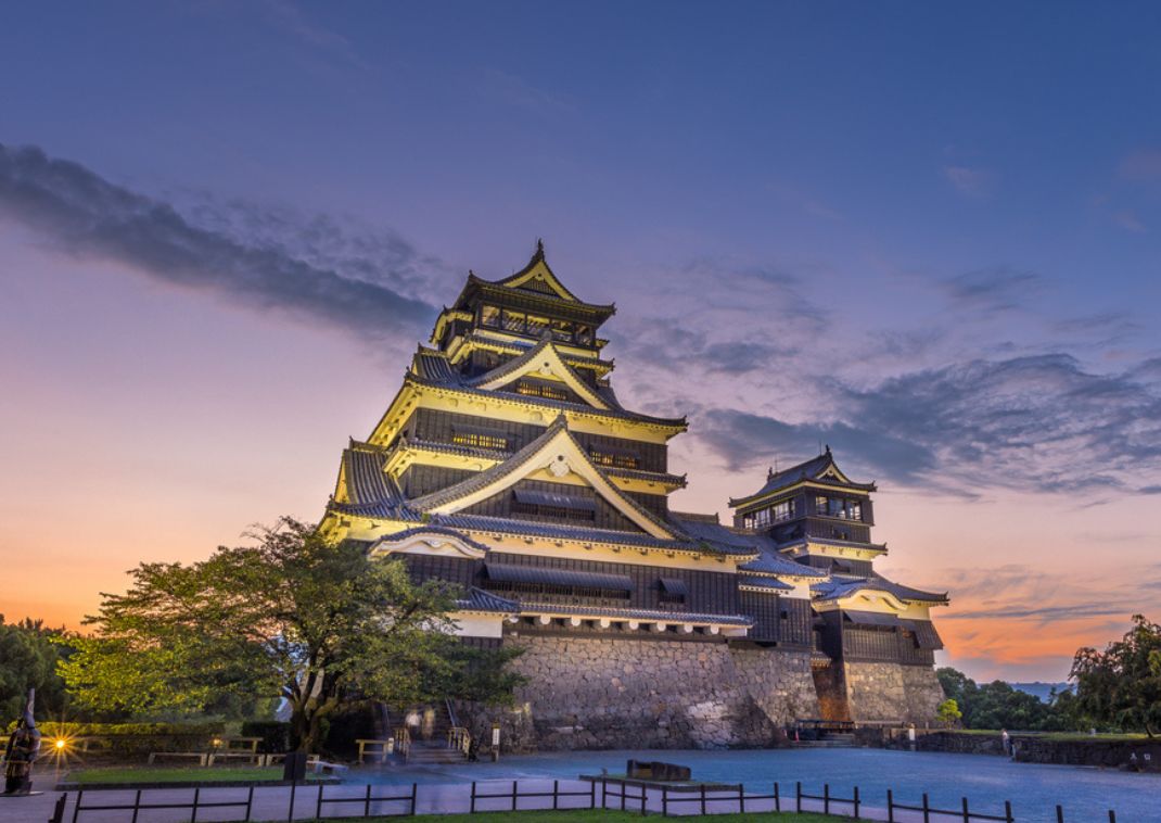 Castillo japonés histórico de paredes de madera negra y varios niveles, iluminado al anochecer con un cielo dramático en tonos púrpura y naranja.