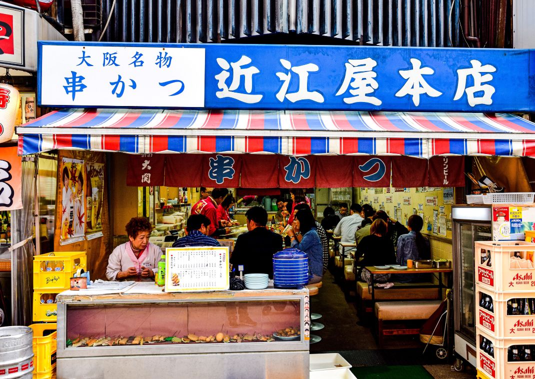 Restaurante japonés concurrido con clientes comiendo en el interior, letrero azul y blanco con caracteres japoneses y cajas de bebidas apiladas afuera.
