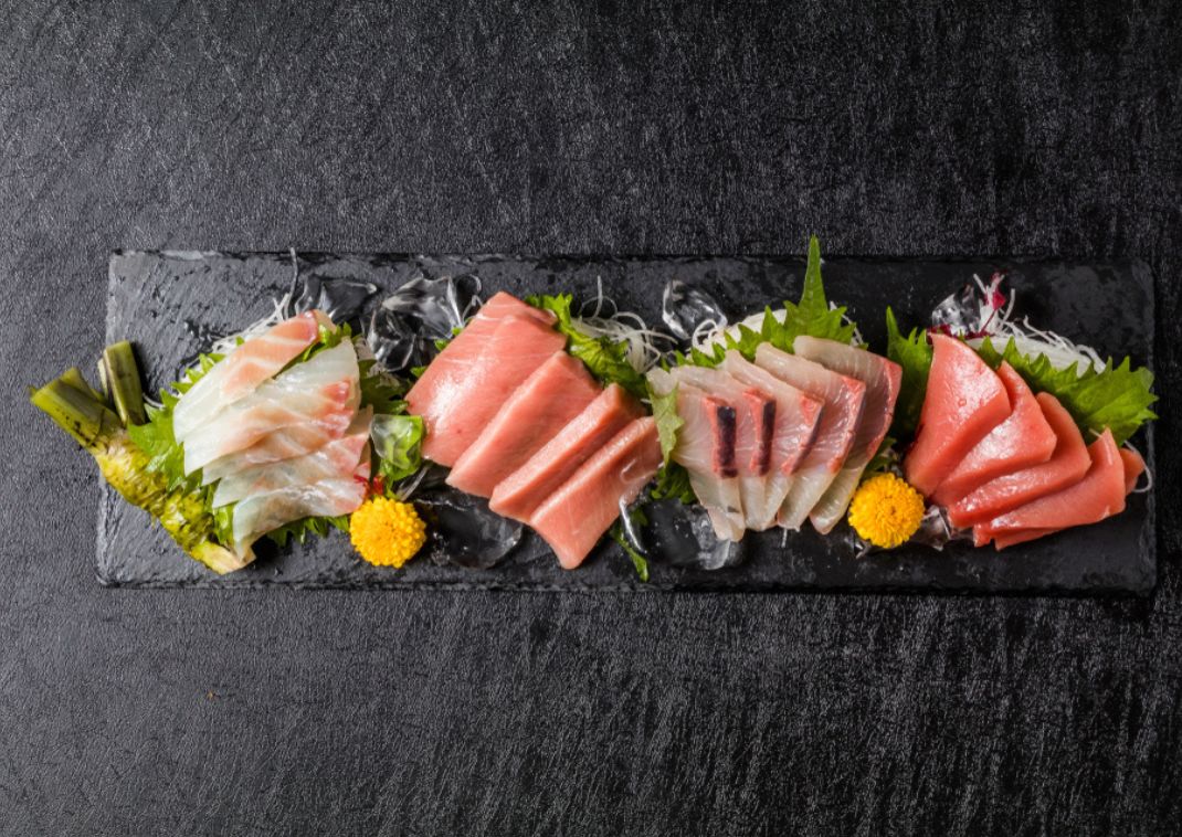  Bandeja de sashimi surtido con láminas de pescado crudo, incluyendo atún y pescado blanco, dispuestas en una pizarra negra con guarnición y flores.