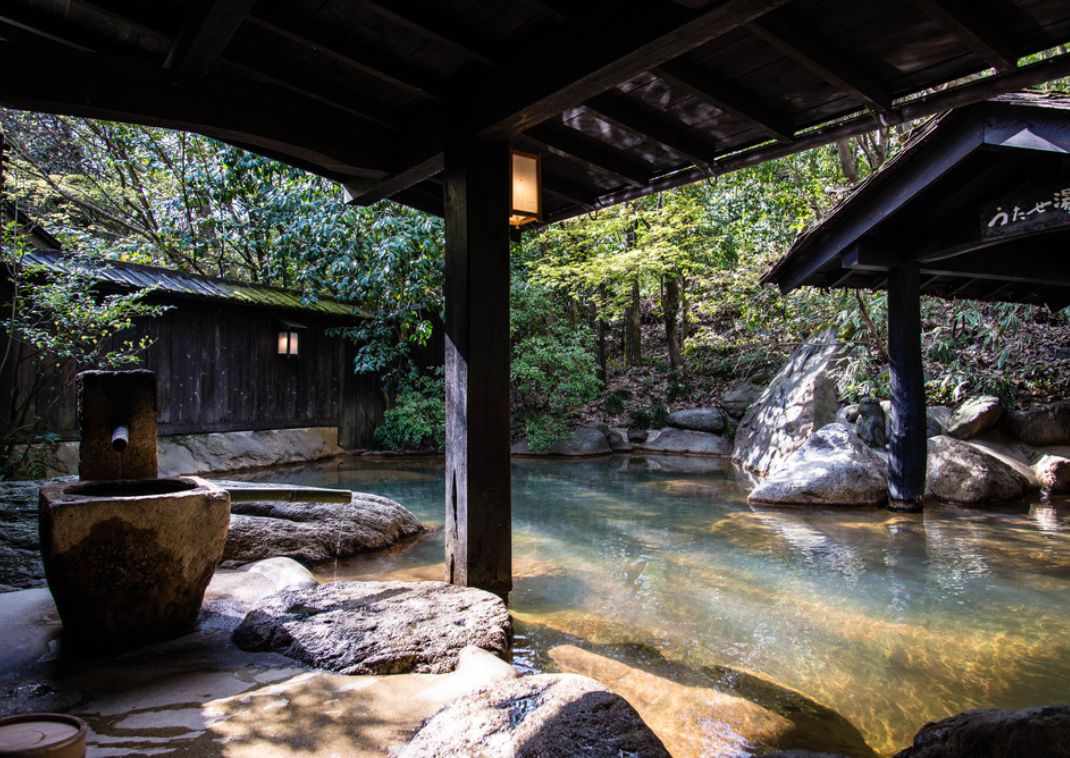 Piscina termal japonesa de color turquesa humeante en un jardín japonés, con puertas torii rojas y árboles densos al fondo.