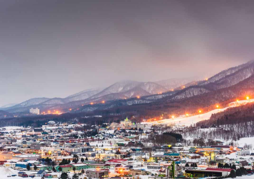 Ciudad nevada y montañas con pistas de esquí iluminadas al fondo
