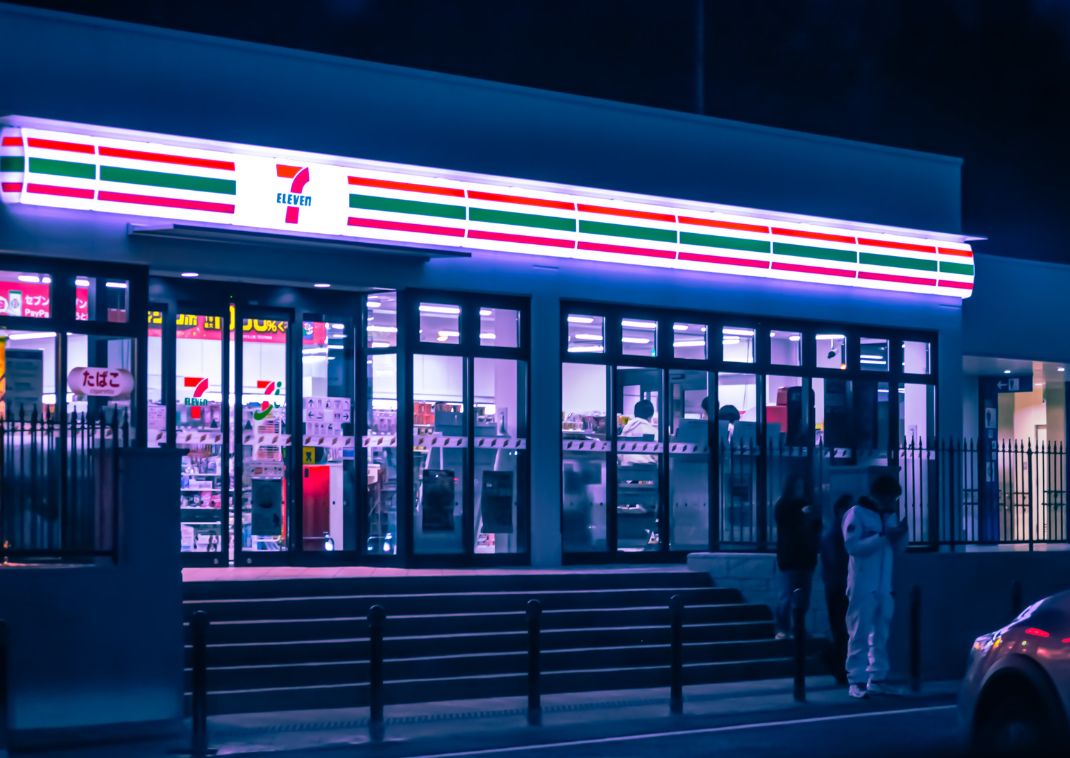 Tienda de conveniencia 7-Eleven en Japón iluminada por la noche, con personas de pie cerca de la entrada.