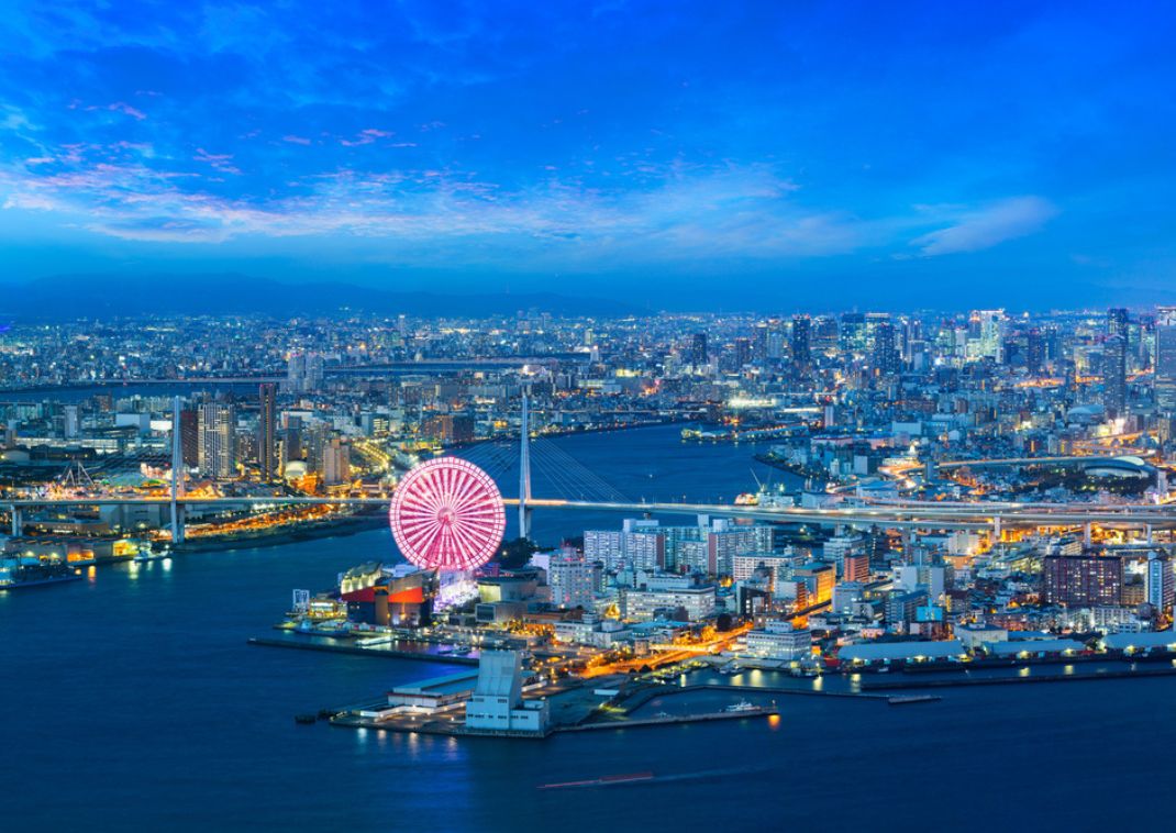 Vista aérea del paseo marítimo de una ciudad al anochecer, con una noria iluminada y edificios encendidos junto al agua