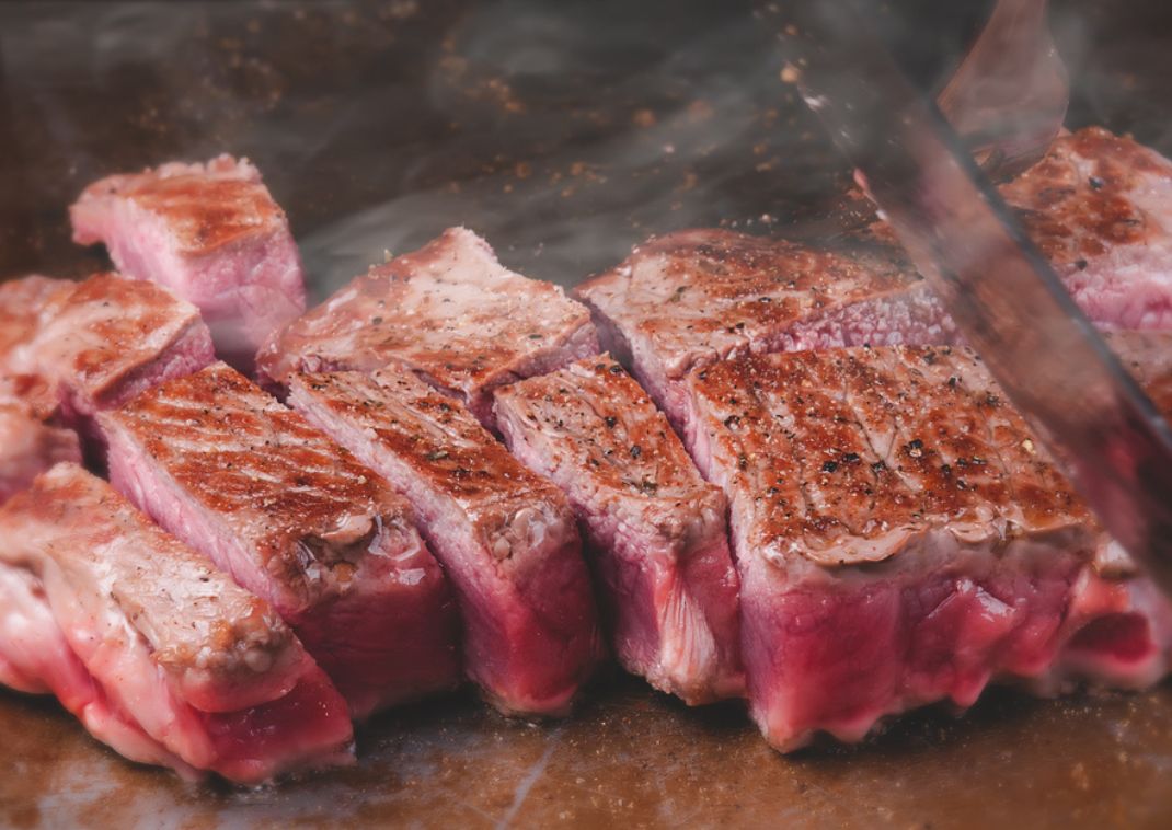 Primer plano de trozos de filete término medio cocinándose en una parrilla caliente, con marcas de la parrilla y vapor visible.