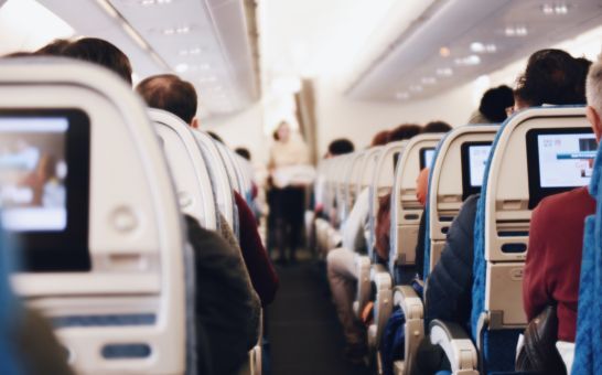 Interior de un gran avión de pasajeros con gente sentada y una azafata de uniforme caminando por el pasillo.