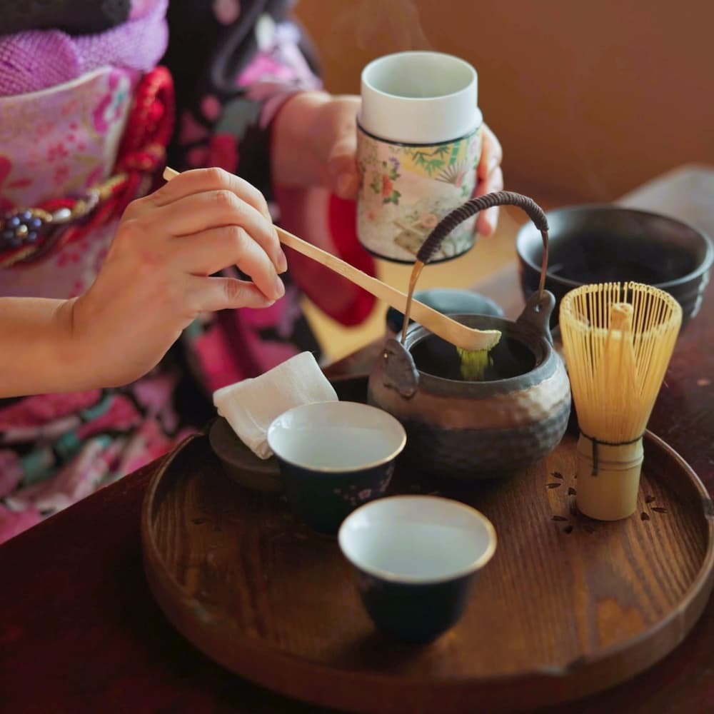 Mujer japonesa preparando té matcha en una ceremonia tradicional del té en casa, con polvo matcha, hierbas y una tetera.