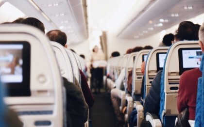 Interior de un gran avión de pasajeros con gente sentada y una azafata de uniforme caminando por el pasillo.